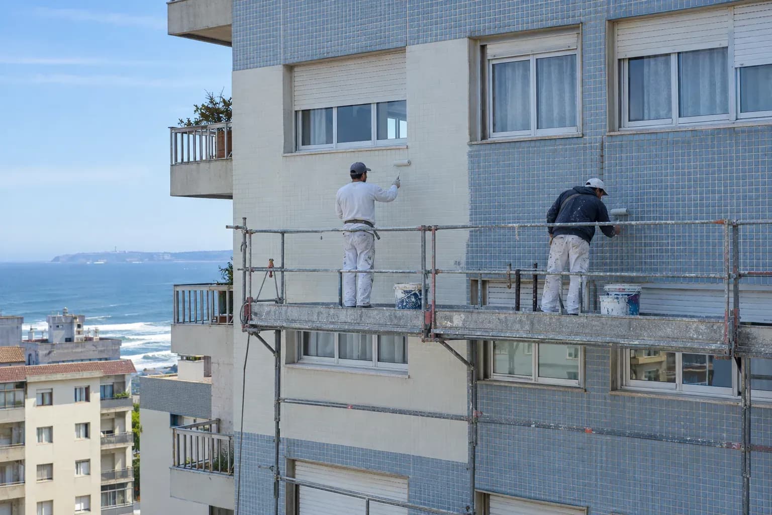 Dois pintores num andaime a pintar a fachada de um prédio com azulejos