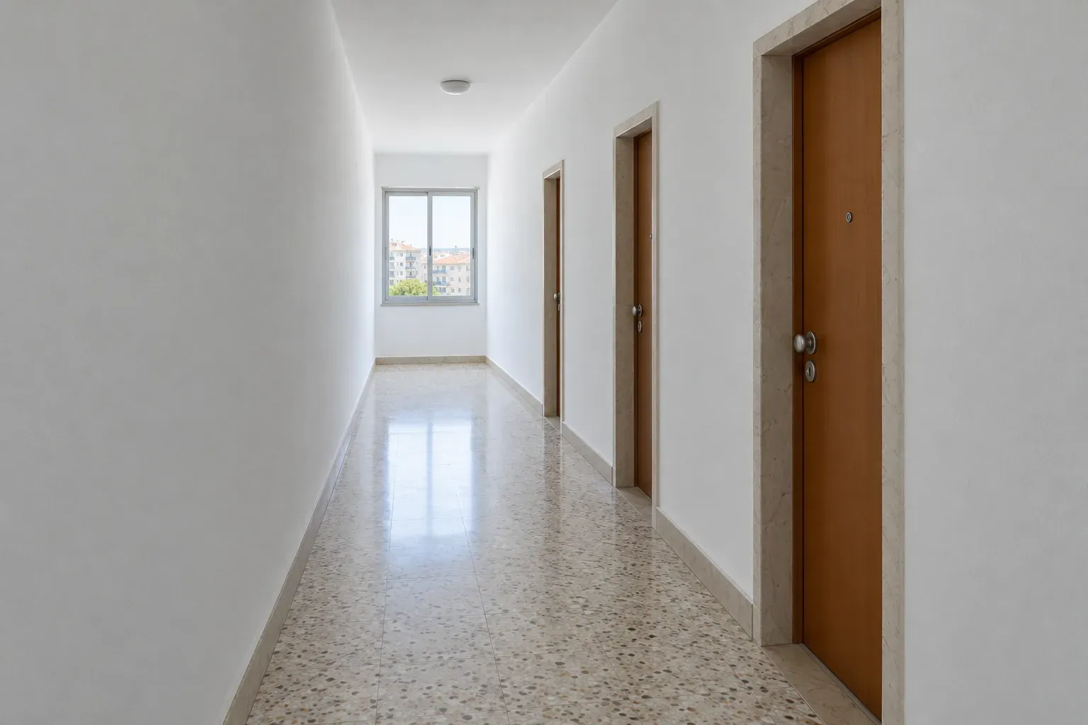 Corredor de um prédio com paredes claras e portas fechadas, com a luz de uma janela ao fundo