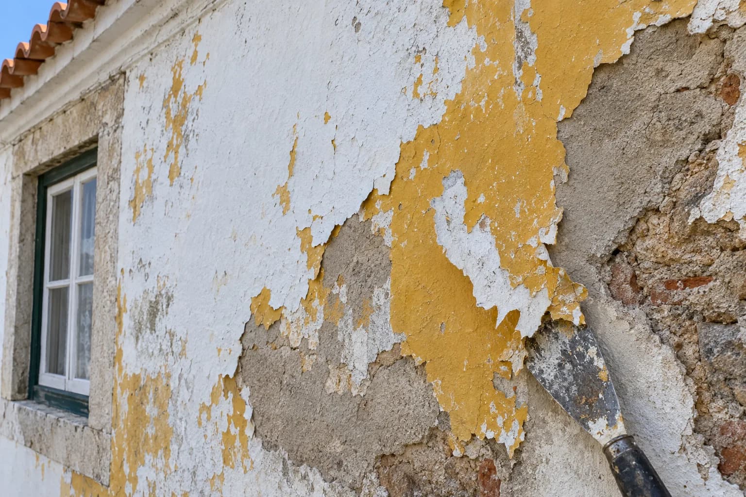 Parede exterior com tinta antiga a descascar e reboco à mostra, com uma espátula encostada